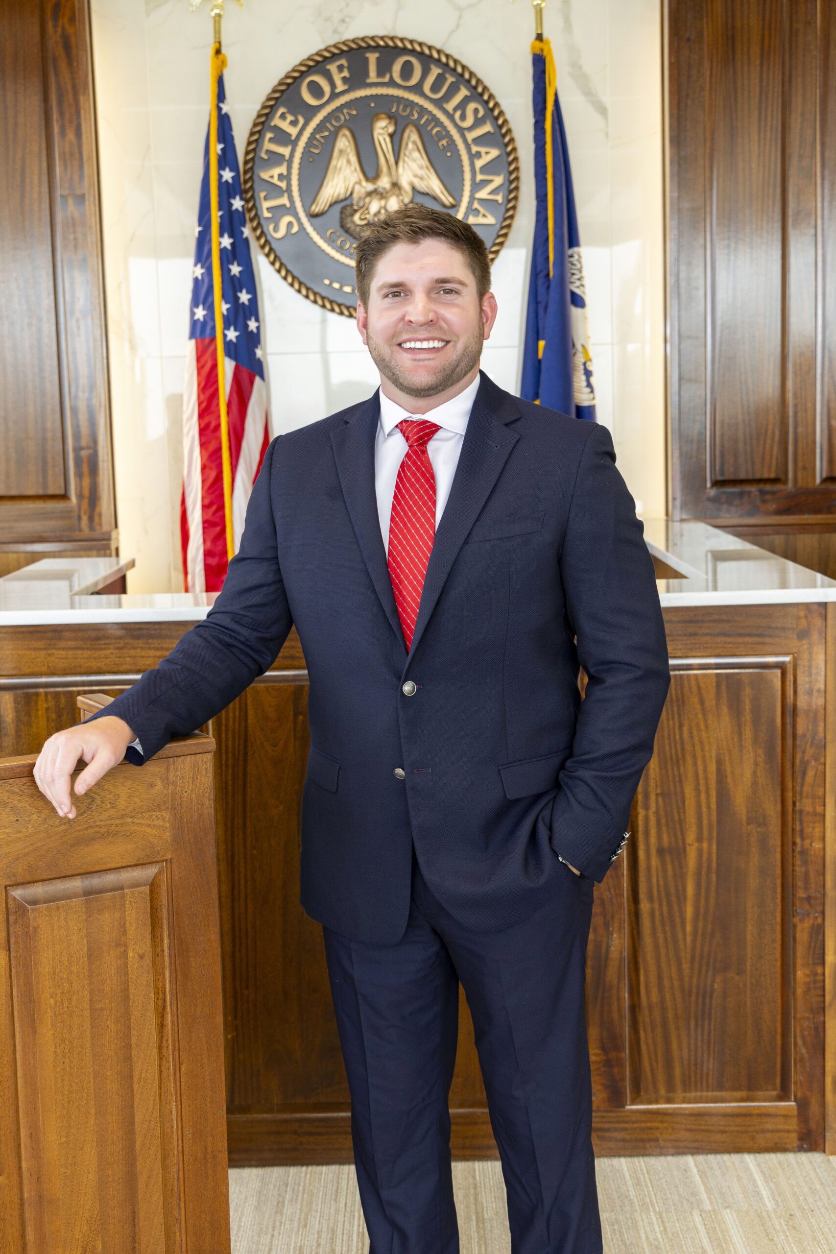 A man in a dark suit and red tie stands before the Louisiana state seal, representing a skilled truck accident lawyer ready to assist.