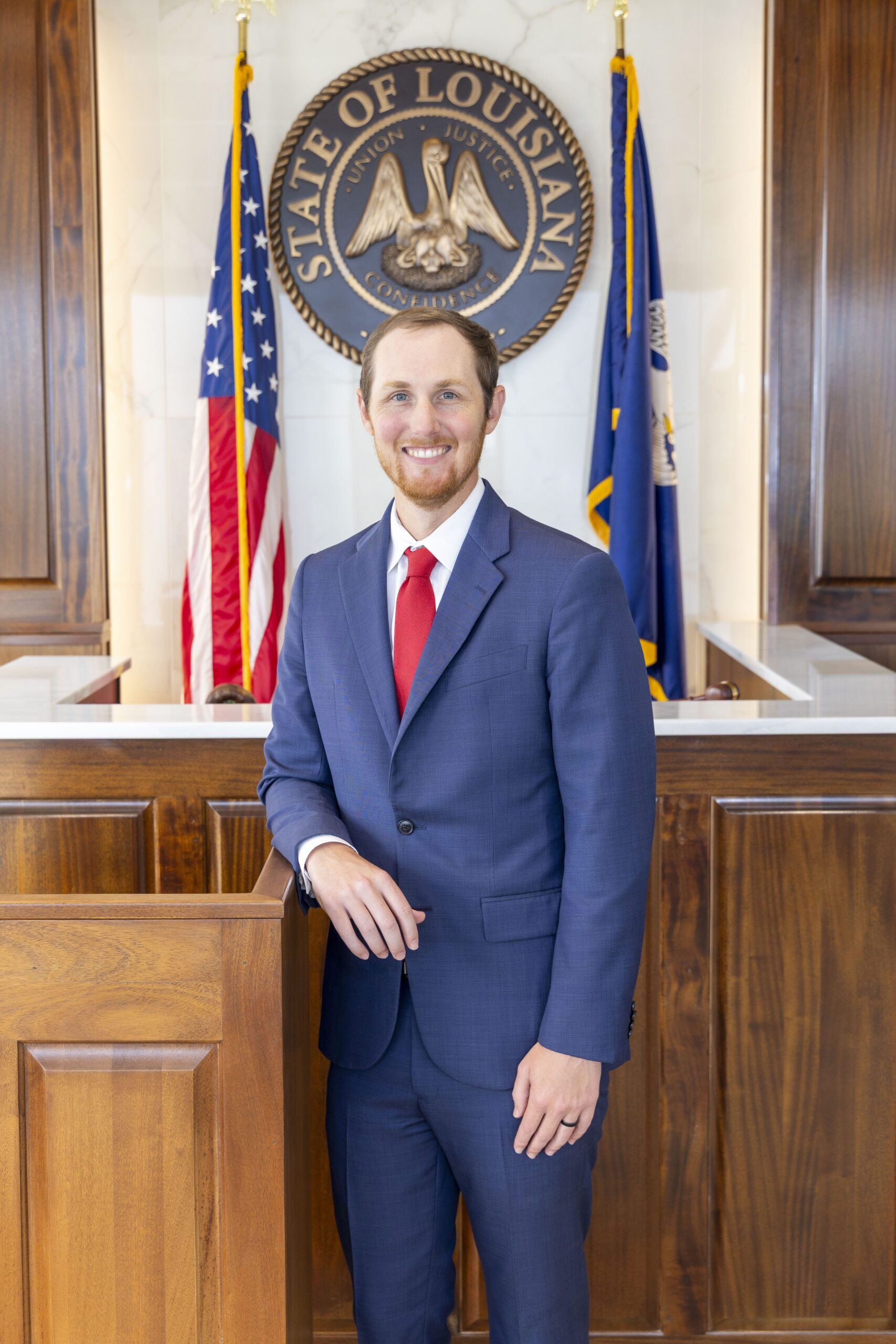 A man in a blue suit and red tie stands in a courtroom with Louisiana and US flags, representing personal injury lawyers serving clients.