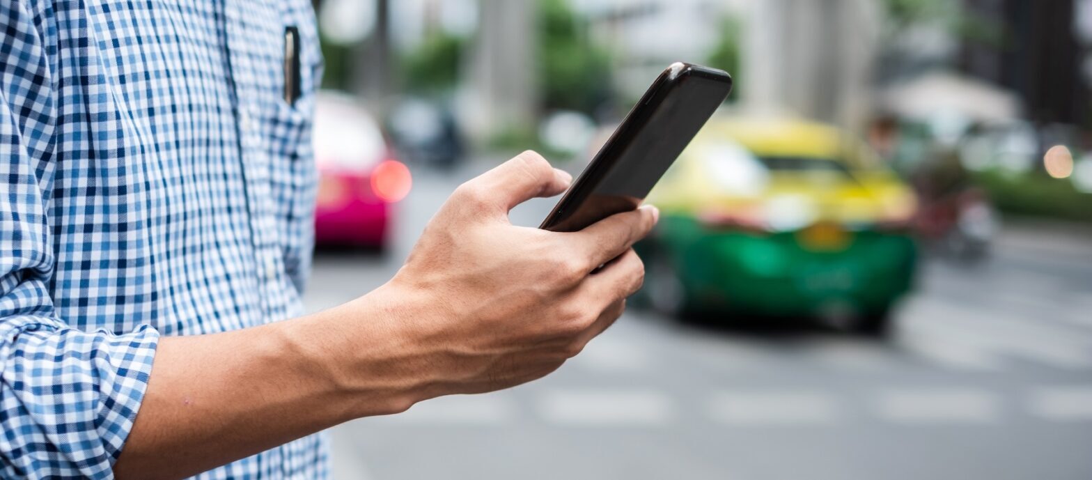 Person in a checkered shirt holds a smartphone on a Baton Rouge street, with blurred cars in the background.